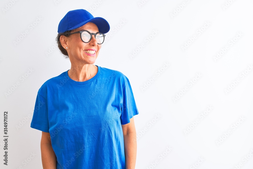 Senior deliverywoman wearing cap and glasses standing over isolated white background looking away to side with smile on face, natural expression. Laughing confident.