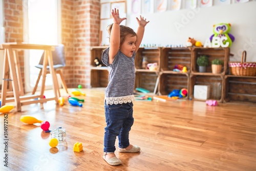 Beautiful toddler standing with hands raised smiling around lots of toys at kindergarten