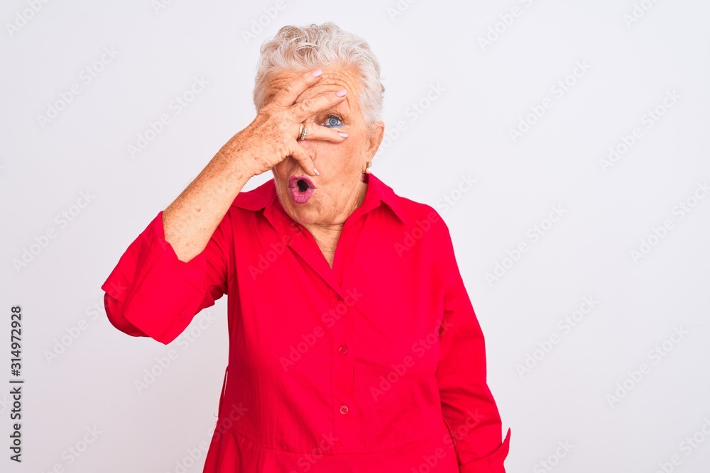 Senior grey-haired woman wearing red casual shirt standing over isolated white background peeking in shock covering face and eyes with hand, looking through fingers with embarrassed expression. Senior grey-haired woman wearing red casual shirt standing over isolated white background peeking in shock covering face and eyes with hand, looking through fingers with embarrassed expression.