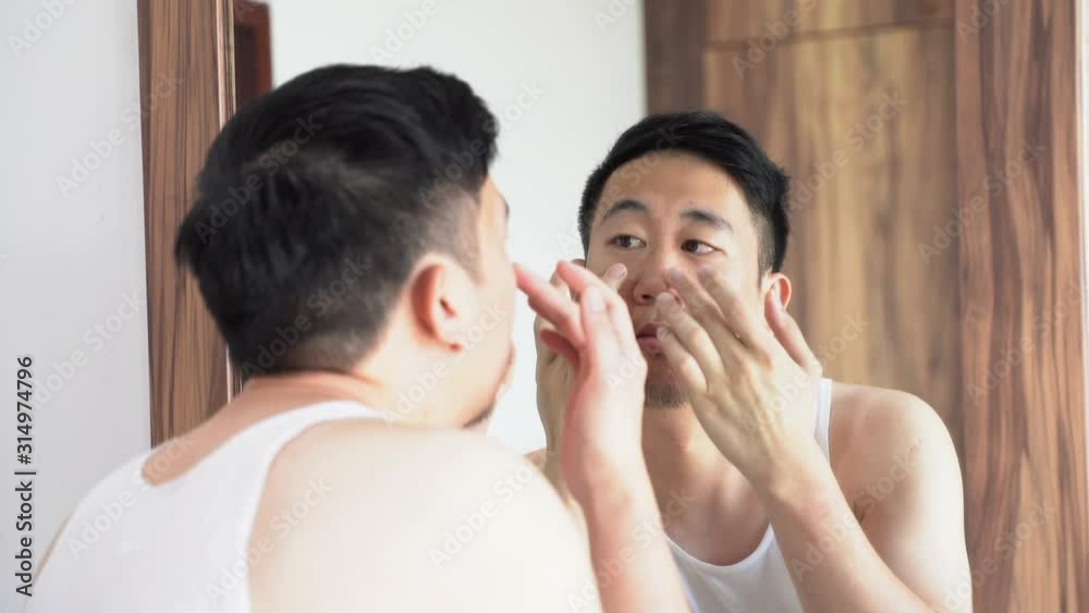 Confused Asian young man in white shirt squeezing pimples in front of mirror at home Confused Asian young man in white shirt squeezing pimples in front of mirror at home