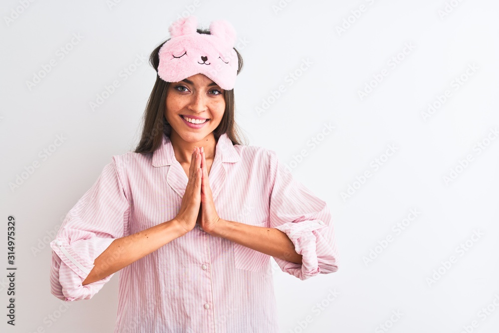 Young beautiful woman wearing sleep mask and pajama over isolated white background praying with hands together asking for forgiveness smiling confident. Young beautiful woman wearing sleep mask and pajama over isolated white background praying with hands together asking for forgiveness smiling confident.