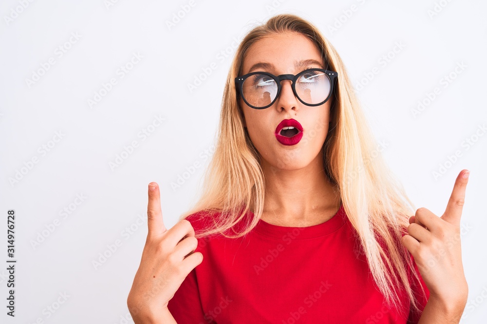Young beautiful woman wearing red t-shirt and glasses standing over isolated white background amazed and surprised looking up and pointing with fingers and raised arms.
