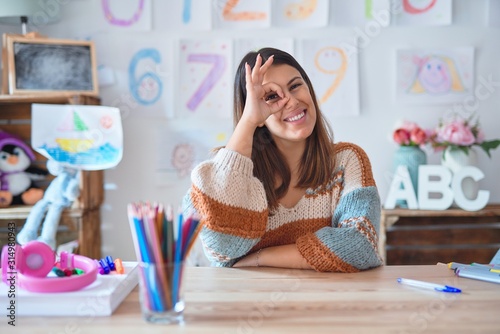 Young beautiful teacher woman wearing sweater and glasses sitting on desk at kindergarten doing ok gesture with hand smiling, eye looking through fingers with happy face.