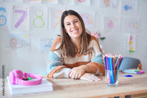 Young beautiful teacher woman wearing sweater and glasses sitting on desk at kindergarten with a happy and cool smile on face. Lucky person.