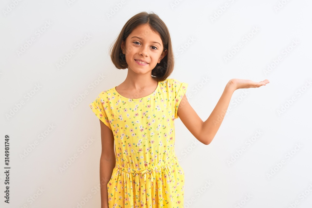Young beautiful child girl wearing yellow floral dress standing over isolated white background smiling cheerful presenting and pointing with palm of hand looking at the camera. Young beautiful child girl wearing yellow floral dress standing over isolated white background smiling cheerful presenting and pointing with palm of hand looking at the camera.
