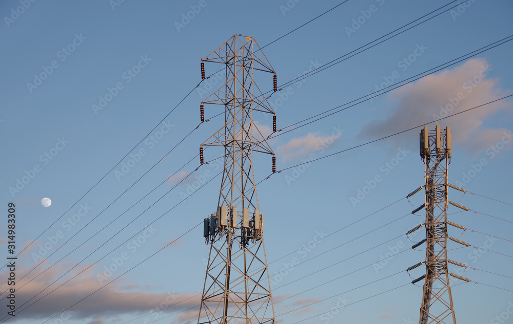 Low angle view of two different electric distribution towers and wires against a blue evening sky with some clouds and the moon visible Low angle view of two different electric distribution towers and wires against a blue evening sky with some clouds and the moon visible