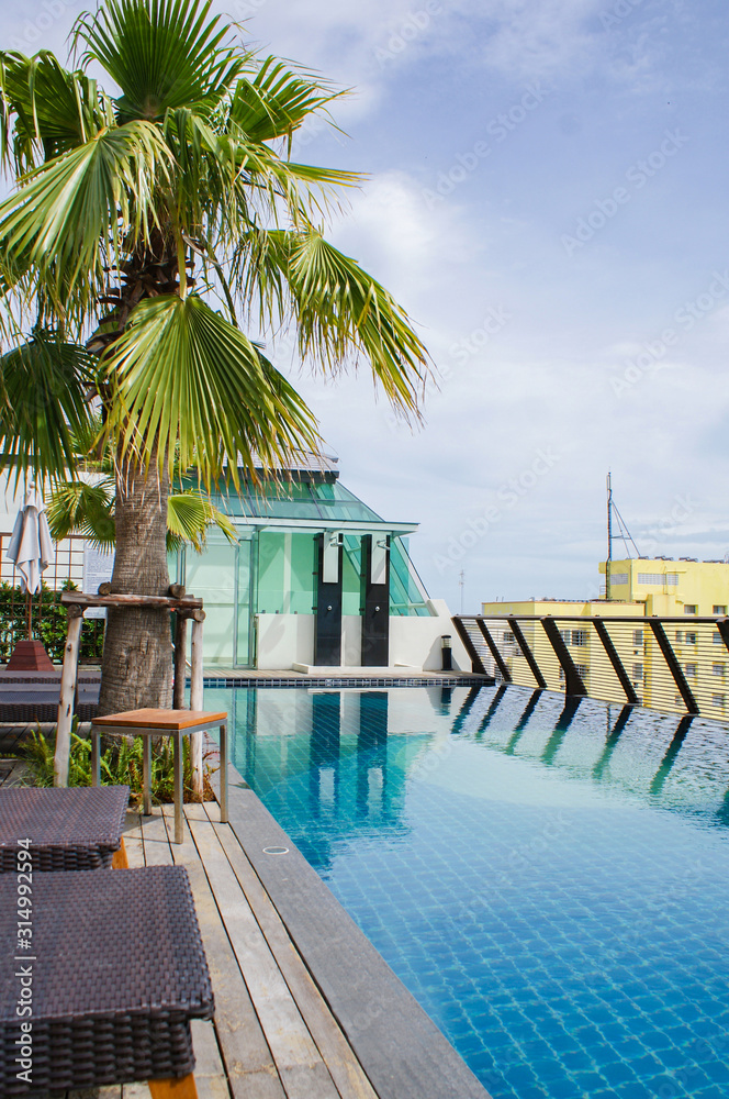 Rooftop pool with sunbeds, a palm tree and showers on a background of blue sky with clouds and a yellow building. Rooftop pool with sunbeds, a palm tree and showers on a background of blue sky with clouds and a yellow building.