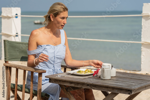 Young woman having romantic breakfast in hotel restaurant during sunrise near sea waves on the tropical beach, close up. Concept of leisure and travel