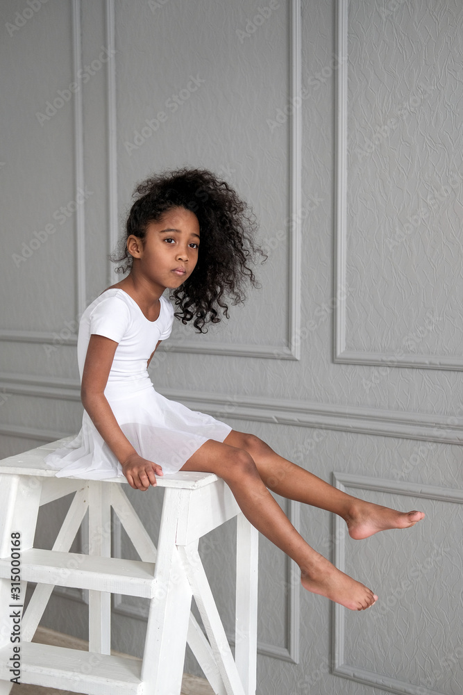 A portrait of an African ballerina in a white gymnastic costume sits on a wooden chair. A portrait of an African ballerina in a white gymnastic costume sits on a wooden chair.