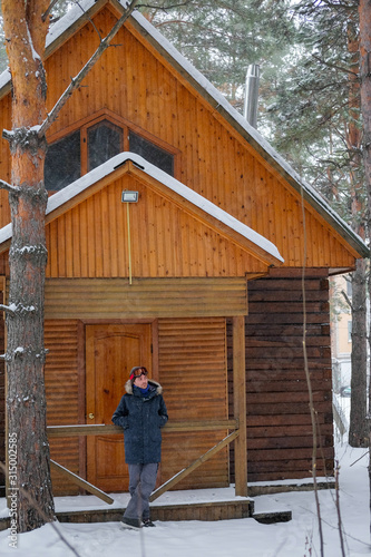Snowboarding man sitting on snowy porch wooden rustic cottage in the winter woods. © evelinphoto