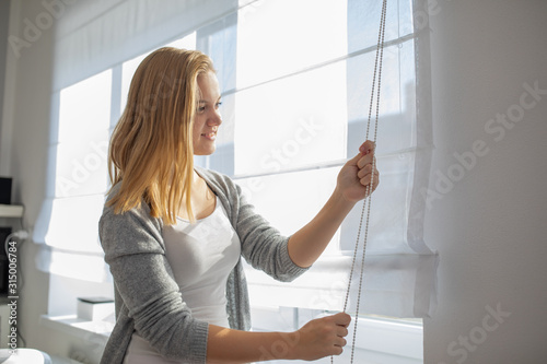 Pretty, young woman lowering the interior shades/blinds in her modern interior apartment photo