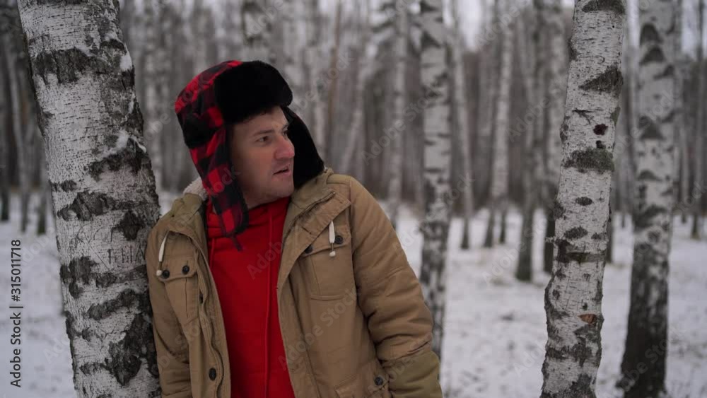 Man standing in forest in winter day Serious young male wearing beige jacket over red hoodie and earflap hat standing on snowy forest and looking away Man standing in forest in winter day Serious young male wearing beige jacket over red hoodie and earflap hat standing on snowy forest and looking away