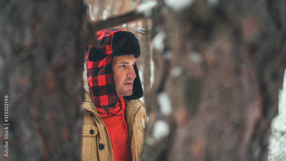 Man standing in forest in winter day. Serious young male wearing beige jacket over red hoodie and earflap hat standing on snowy forest and looking away Man standing in forest in winter day. Serious young male wearing beige jacket over red hoodie and earflap hat standing on snowy forest and looking away