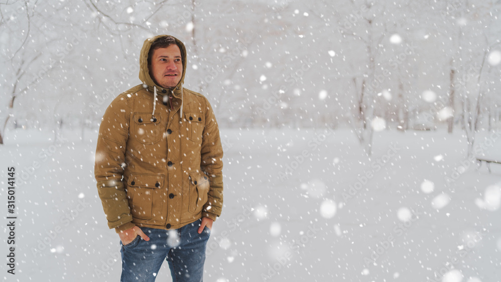 Portrait of young man in jacket and jeans in winter season. Handsome man stands and squint in winter park. Fluffy snow envelops everything around. Portrait of young man in jacket and jeans in winter season. Handsome man stands and squint in winter park. Fluffy snow envelops everything around.