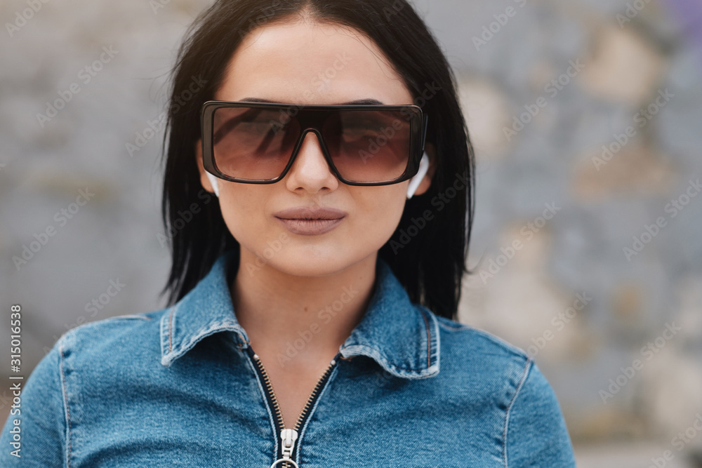 One Beautiful Brunette Girl Dressed in Denim Overalls Uses Mobile Bluetooth Earphones One Beautiful Brunette Girl Dressed in Denim Overalls Uses Mobile Bluetooth Earphones