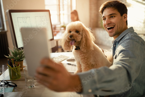 Happy freelance worker using touchpad while taking selfie with his dog in the office.
