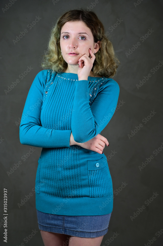 Portrait of a pretty student girl with beautiful curly hair is standing in front of the camera on a gray background. Concept vertical photo of a young woman with emotions in a blue dress. Portrait of a pretty student girl with beautiful curly hair is standing in front of the camera on a gray background. Concept vertical photo of a young woman with emotions in a blue dress.