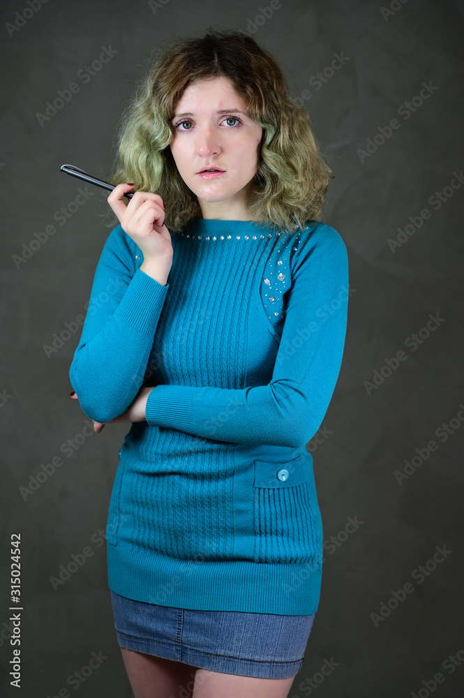 Fototapeta premium Concept vertical photo of a young woman with emotions in a blue dress. Portrait of a pretty student girl with beautiful curly hair is standing in front of the camera on a gray background.