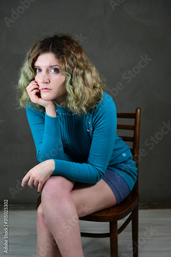 Concept vertical photo of a young woman in a blue dress sitting on a chair. Portrait of a pretty student girl with beautiful curly hair in front of the camera on a gray background.