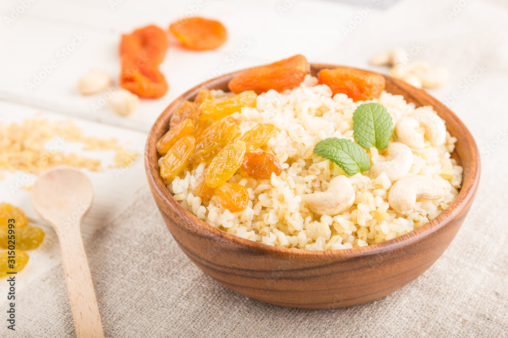 Bulgur porridge with dried apricots, raisins and cashew in wooden bowl on a white wooden background and linen textile. Side view, selective focus. Bulgur porridge with dried apricots, raisins and cashew in wooden bowl on a white wooden background and linen textile. Side view, selective focus.