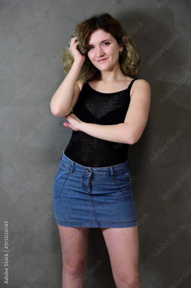 Concept vertical photo of a young woman in a black T-shirt and blue skirt standing straight. Portrait of a pretty student girl with beautiful curly hair in front of the camera on a gray background.