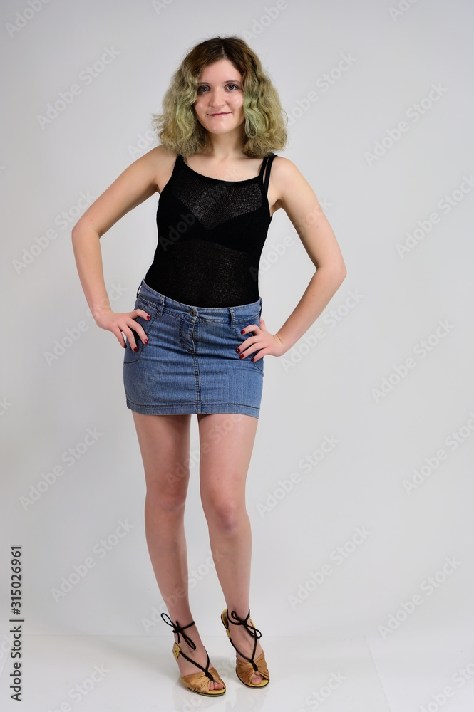 A horizontal photo concept of a young woman in a black t-shirt and blue skirt is standing in front of the camera on a white background. Full-length portrait of a pretty girl with beautiful curly hair. A horizontal photo concept of a young woman in a black t-shirt and blue skirt is standing in front of the camera on a white background. Full-length portrait of a pretty girl with beautiful curly hair.