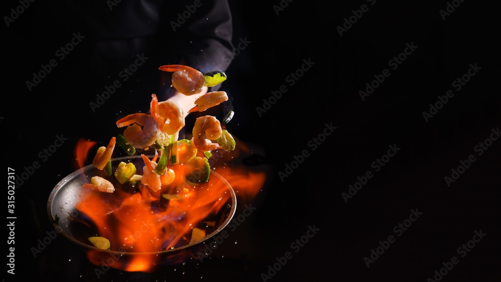 Professional cook cooks shrimps with vegetables on fire. Cooking seafood, healthy vegetarian food, roasting over an open fire. on a dark background. Hotel service, oriental cuisine asia food