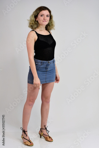 A horizontal photo concept of a young woman in a black t-shirt and blue skirt is standing in front of the camera on a white background. Full-length portrait of a pretty girl with beautiful curly hair. © Вячеслав Чичаев