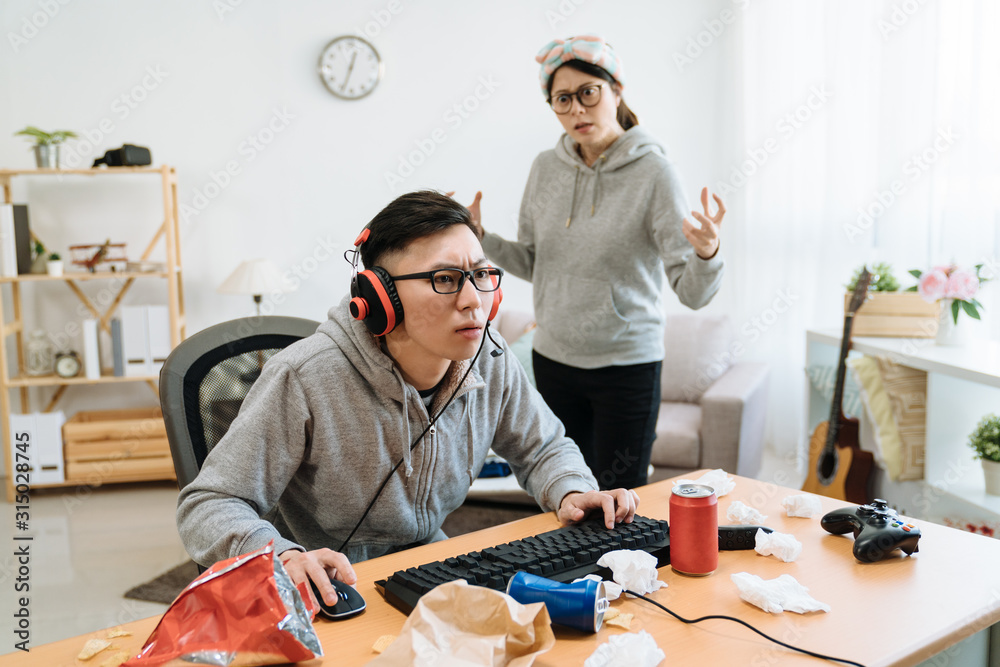 Asian Young Girl Friend Angry At Boyfriend Playing Online Game On Computer.  Mad Female Pushing Guy And Scolding At Him. Frowning Man Ignores His  Annoyed Lover And Wearing Headphones Keep Having Fun.