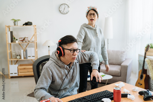 Asian Young Girl Friend Angry At Boyfriend Playing Online Game On Computer.  Mad Female Pushing Guy And Scolding At Him. Frowning Man Ignores His  Annoyed Lover And Wearing Headphones Keep Having Fun.