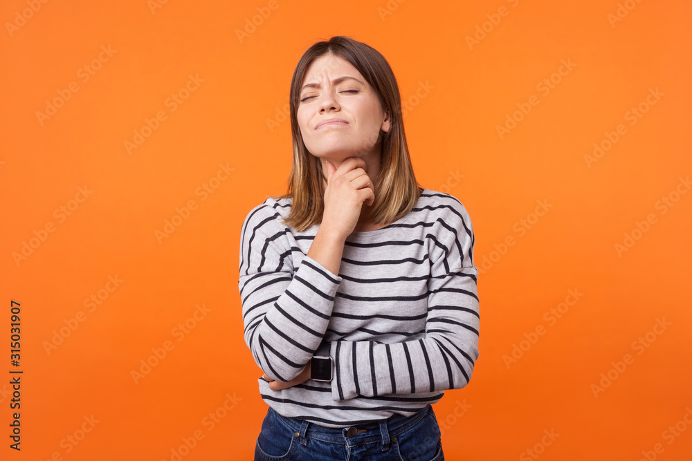Portrait of ill woman with brown hair in long sleeve striped shirt standing touching neck and frowning from pain, suffering sore throat, flu symptom. indoor studio shot isolated on orange background Portrait of ill woman with brown hair in long sleeve striped shirt standing touching neck and frowning from pain, suffering sore throat, flu symptom. indoor studio shot isolated on orange background