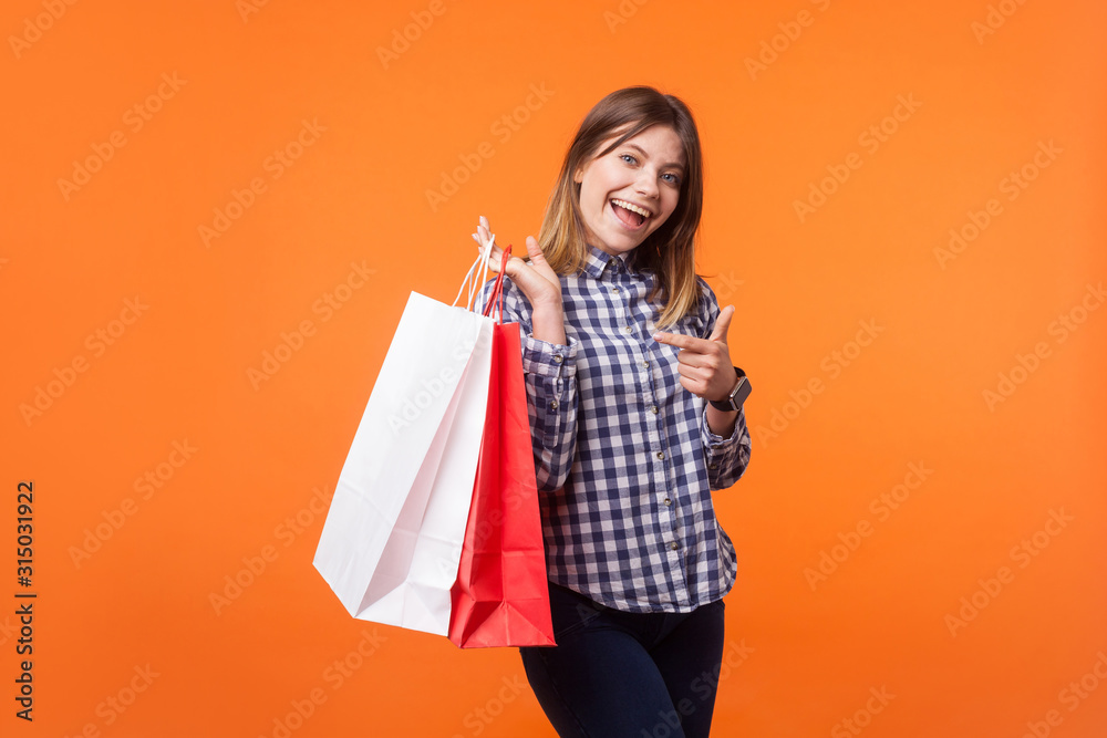 Portrait of lovely brunette woman with charming smile in casual checkered shirt standing pointing at shopping bags, looking satisfied with purchase. indoor studio shot isolated on orange background
