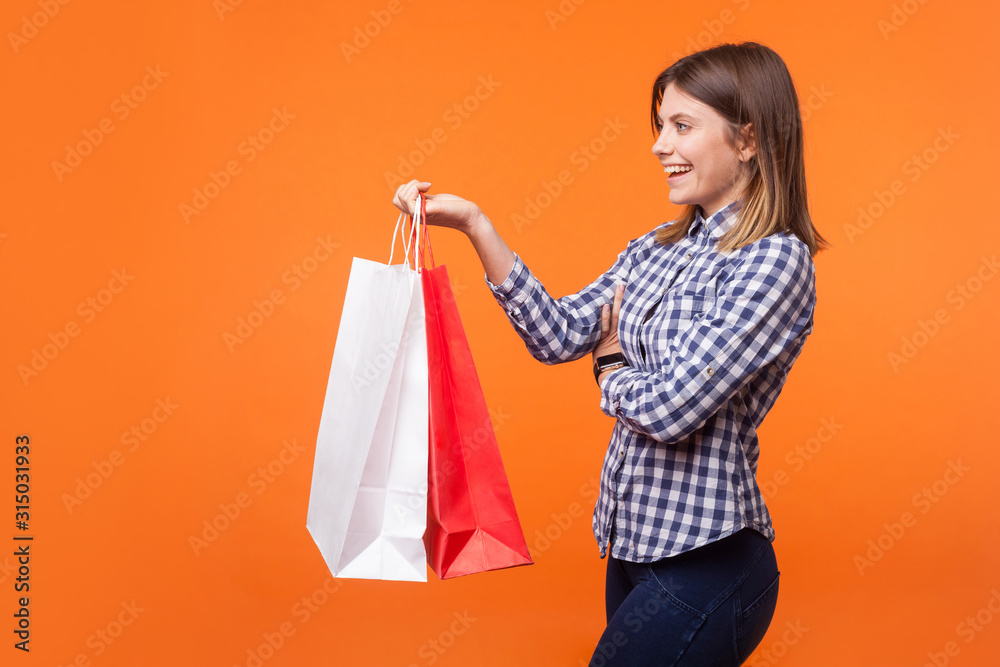 Side view portrait of friendly brunette woman with charming smile in casual checkered shirt standing giving shopping bags and smiling, offering goods. indoor studio shot isolated on orange background