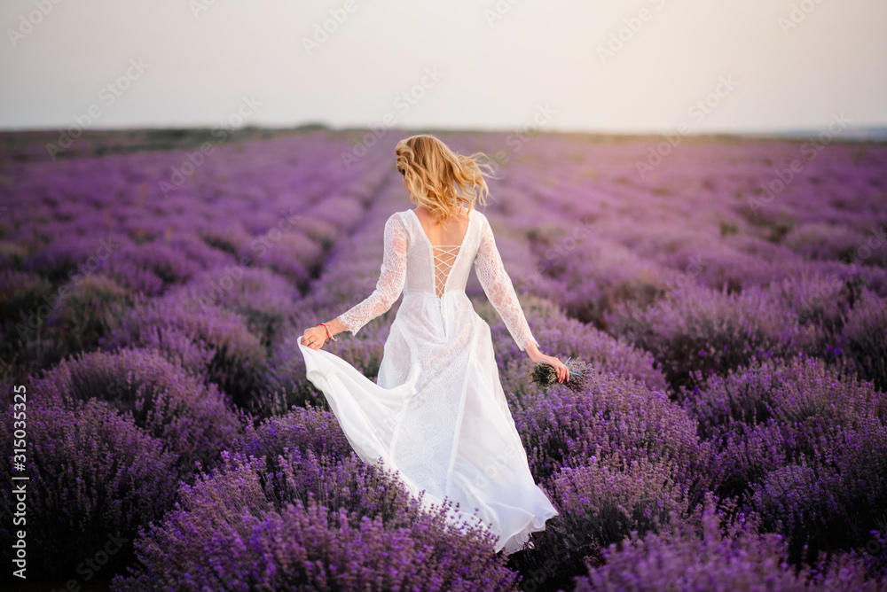 Young woman in white dress walks through blooming lavender field at sunset