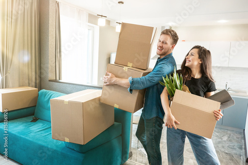 young smiling couple husband and wife looking happy standing with boxes in their hands in new appartment unpacking boxes