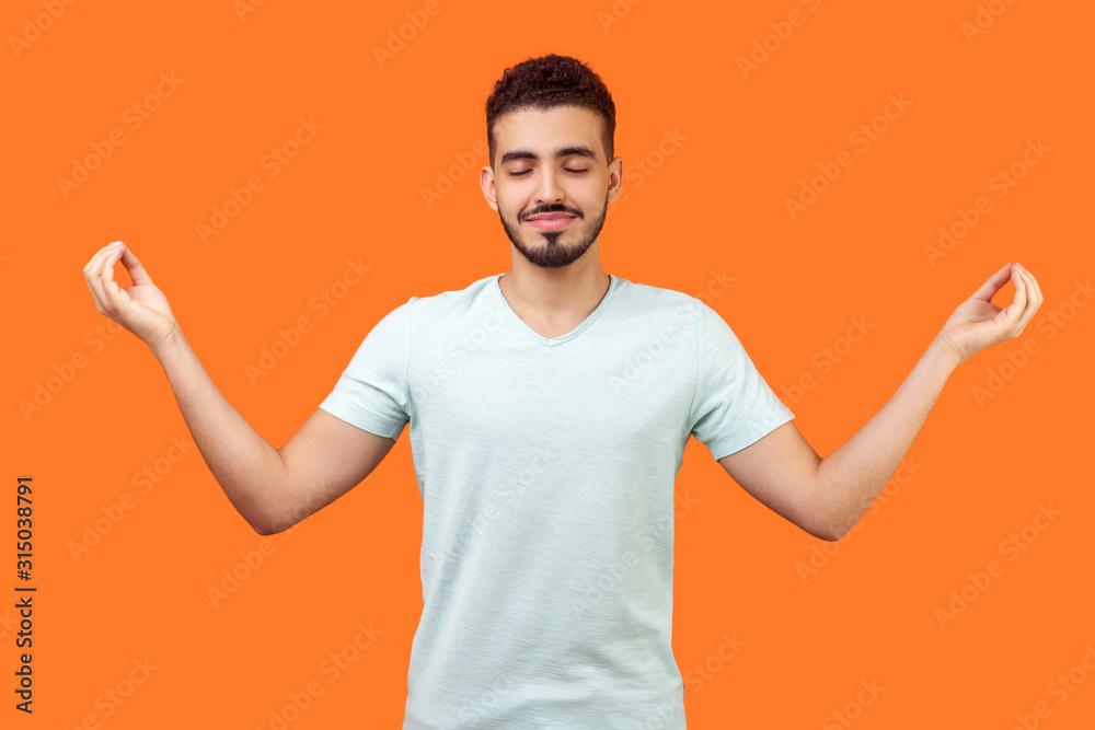 Practice mindfulness. Portrait of peaceful handsome brunette man with beard in white t-shirt doing meditation gesture with fingers, managing stress. indoor studio shot isolated on orange background Practice mindfulness. Portrait of peaceful handsome brunette man with beard in white t-shirt doing meditation gesture with fingers, managing stress. indoor studio shot isolated on orange background