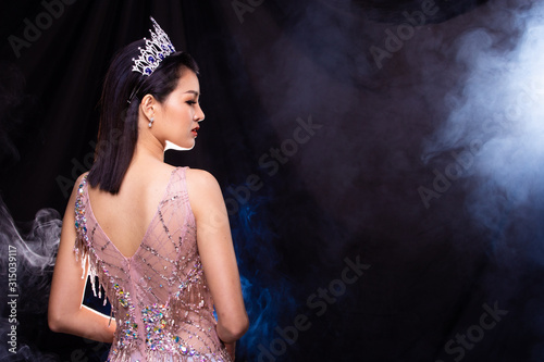 Portrait of Miss Pageant Beauty Contest in Pink sequin Evening Ball Gown with Diamond Crown, Asian Woman points waves hand to side over dark background smoke copy space photo