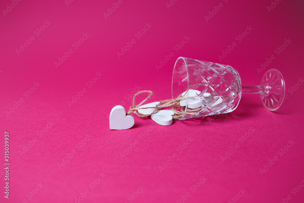 Small white wooden hearts in the lying wine glass on pink background with copy space. Small white wooden hearts in the lying wine glass on pink background with copy space.