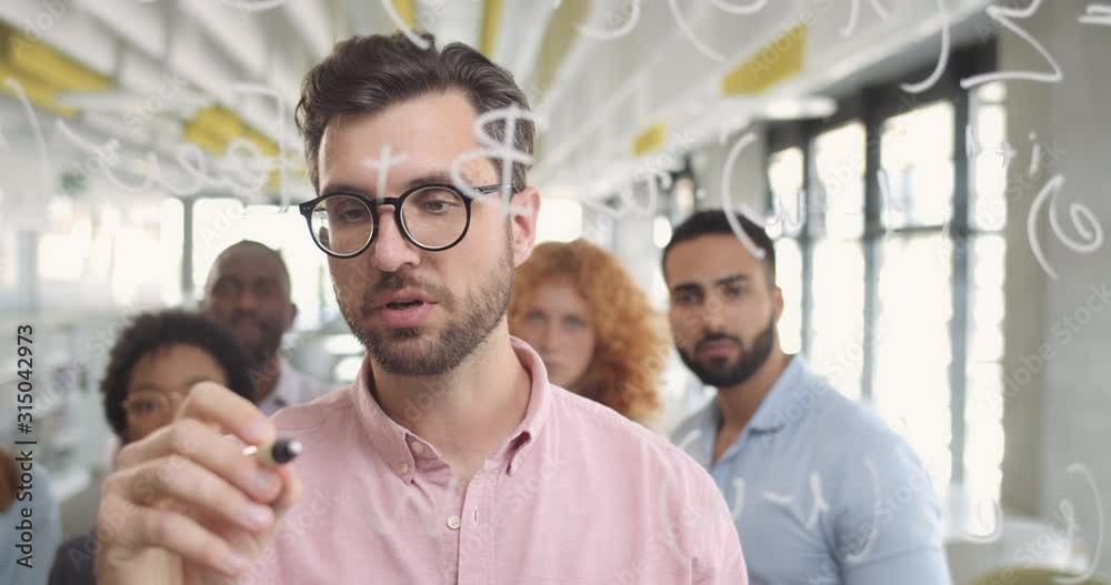 Male office worker in glasses writing on glass board and explaining concept to team members. Close up of handsome bearded man standing near glass wall at corporate conference.