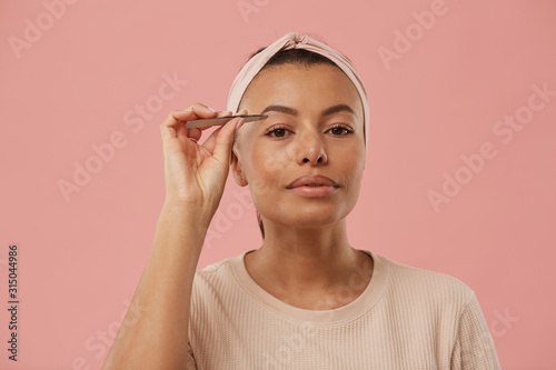 Head and shoulders portrait of beautiful mixed race woman plucking eyebrows while putting on makeup in morning standing against dust pink background, copy space