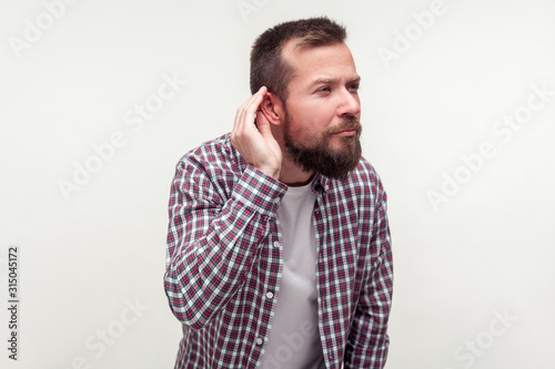 Portrait of deaf bearded man in plaid shirt standing with hand near ear and listening carefully to secret conversation, hearing problems, deafness. indoor studio shot isolated on white background