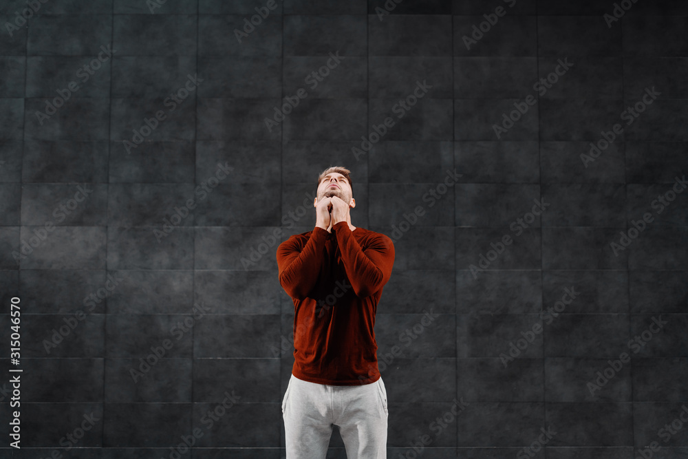 Attractive fit caucasian breaded sportsman in tracksuit doing warm up exercises for neck before running. In background is gray wall. - obrazy, fototapety, plakaty 