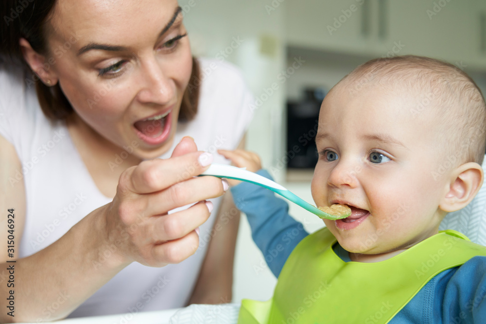 Smiling 8 month Old Baby Boy At Home In High Chair Being Fed Solid Food By Mother With Spoon Smiling 8 month Old Baby Boy At Home In High Chair Being Fed Solid Food By Mother With Spoon