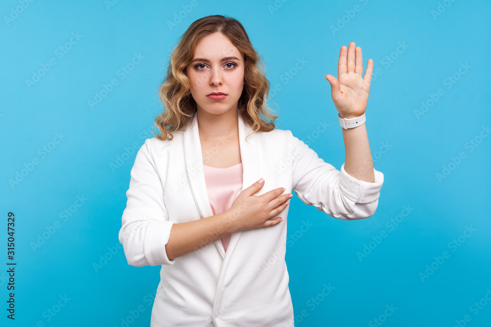 I promise. Portrait of beautiful woman with wavy hair in white jacket making swearing gesture and holding arm on chest, taking oath, pledging allegiance. indoor studio shot isolated on blue background