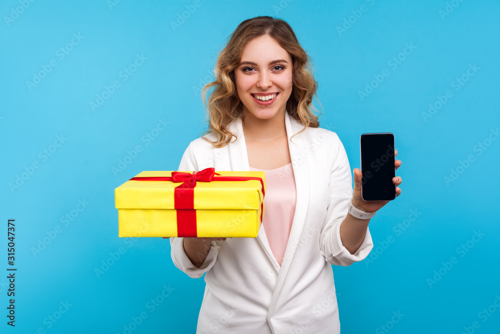 Gift, bonuses in mobile tariff. Portrait of cheerful woman with wavy hair in white jacket showing wrapped present box and cell phone, smiling at camera. indoor studio shot isolated on blue background Gift, bonuses in mobile tariff. Portrait of cheerful woman with wavy hair in white jacket showing wrapped present box and cell phone, smiling at camera. indoor studio shot isolated on blue background