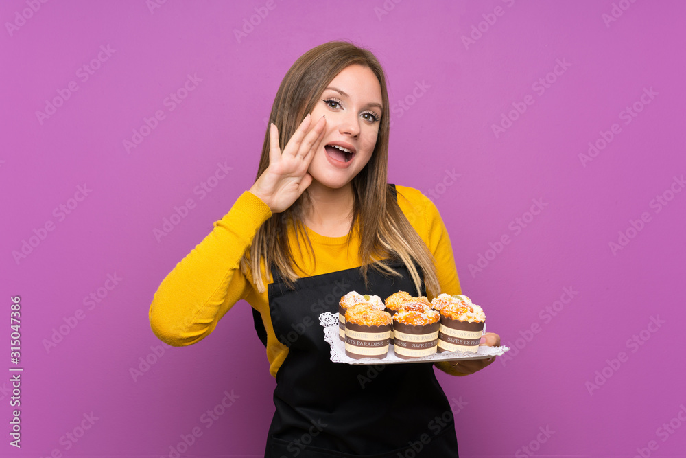 Teenager girl holding lots of different mini cakes over isolated purple background shouting with mouth wide open Teenager girl holding lots of different mini cakes over isolated purple background shouting with mouth wide open