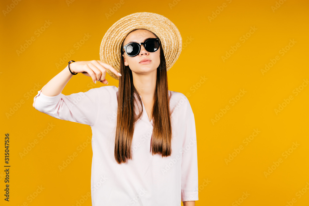 pointing finger shows down. girl stands in a hat and black glasses on a yellow background pointing finger shows down. girl stands in a hat and black glasses on a yellow background