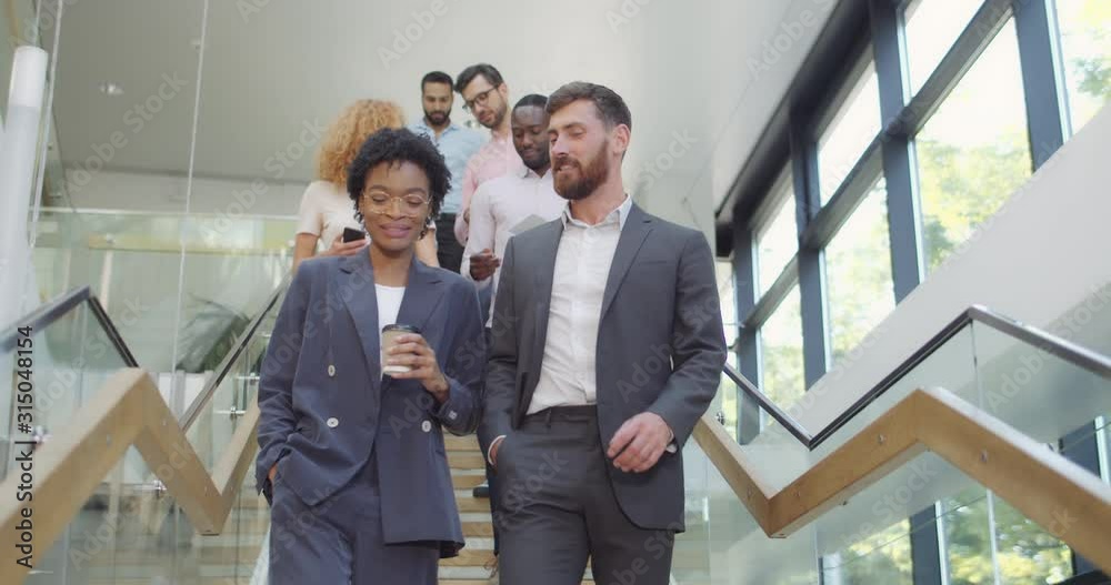 Multi ethnic office workers walking downstairs smiling and communicating after corporate meeting. Group of male and female employees descending modern office stairs and talking.