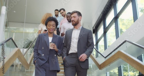 Multi ethnic office workers walking downstairs smiling and communicating after corporate meeting. Group of male and female employees descending modern office stairs and talking.