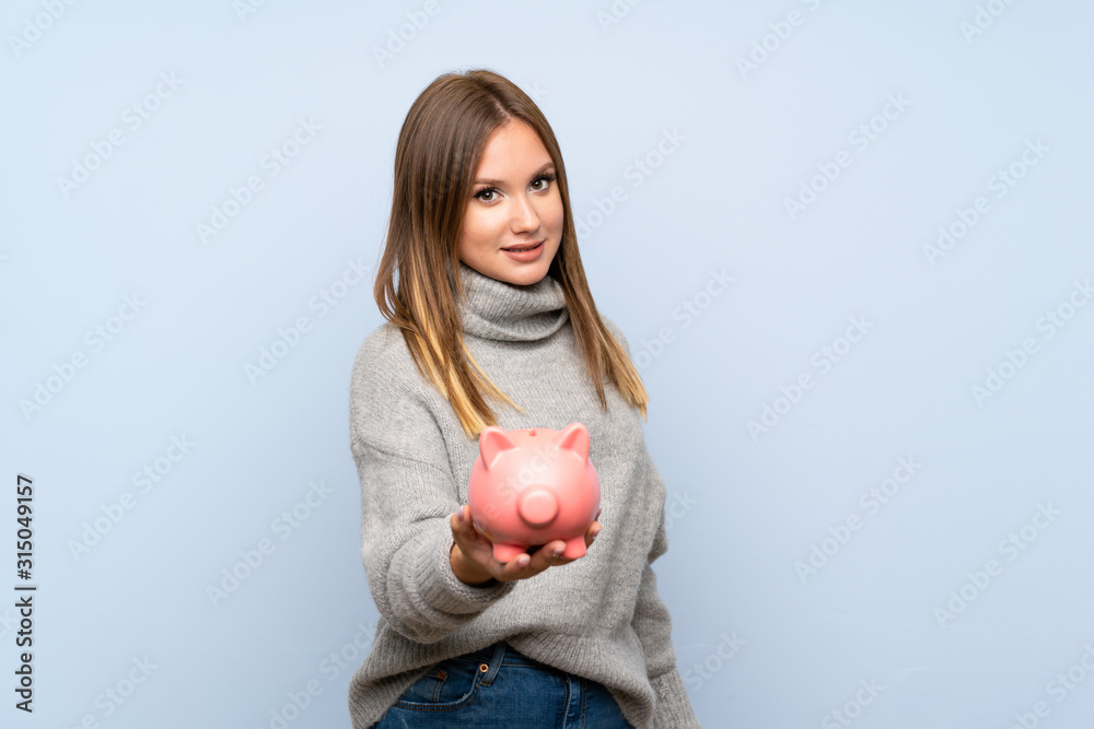 Teenager girl with sweater over isolated blue background holding a big piggybank Teenager girl with sweater over isolated blue background holding a big piggybank